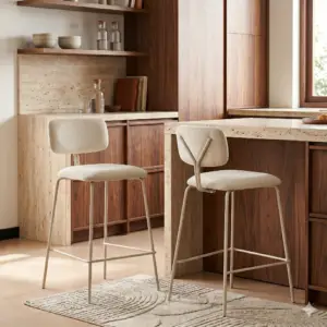 שני כסאות בר בצבע שמנת מדגם BAR ARSENAL, בעלי רגלי מתכת מעוצבות, ניצבים ליד אי מטבח עם משטח אבן, מוקפים בארונות עץ, מדפים פתוחים שעליהם מוצגים כלי אוכל, ושטיח בז' מרקם מתחת.