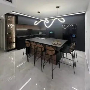 מטבח מודרני עם אי מרכזי הכולל משטח אפור ושישה כסאות בר גבוהים BAR JIN עם גב. אור התליון הגלי המסוגנן מאיר את החלל, משלים על ידי ארונות בגוון כהה ורצפת אריחים לבנה מבריקה.