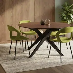 שולחן אוכל עגול מעץ עם רגליים שחורות מצטלבות, המוקף בארבעה כיסאות מרופדים בירוק, ניצב על שטיח בעל מרקם בחדר מודרני עם קירות מחופים עץ ועציץ בפינה.