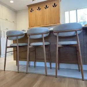 שלושה כיסאות בר מודרניים BAR KALI PU עם רגלי עץ ומושבים מרופדים באפור בהיר מסודרים בשורה מול אי מטבח מעץ במטבח מואר ועכשווי עם פרזול עץ ולבן.