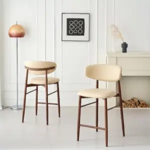 שני כסאות בר מדגם BAR KALI (בדמוי עור ובגוון אגוז) ניצבים בחדר מואר ומודרני, עם קירות מחופים בלוחות עץ לבנים, מנורת רצפה, יצירות אמנות מינימליסטיות, עשב מיובש באגרטל ועצי הסקה מתחת לאח.