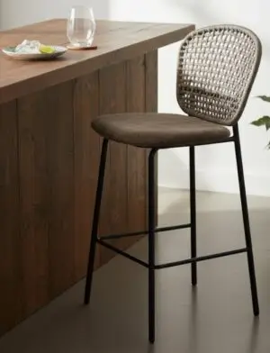 כיסא בר "A BAR KALI" (בד דמוי עור ובגוון אגוז) עם משענת גב קלועה ומושב מרופד ניצב ליד דלפק עץ שעליו מונחים כוס מים, חטיפים בצלחת ופרוסת ליים.