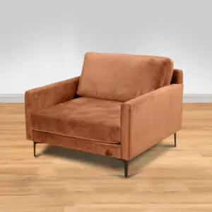 ARM CHAIR MAMBA הוא כורסה רחבה ומודרנית עם ריפוד קטיפה בצבע חלודה, רגלי מתכת דקיקות וקווים נקיים, המוצבת על רצפת עץ בהירה על רקע אפור-לבן אחיד.
